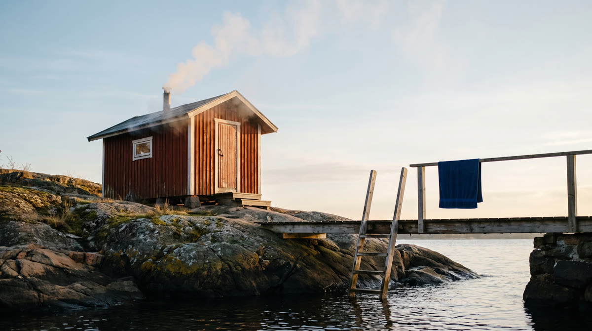 Red wooden sauna on a rocky shore by the pier, blue towel on the railing